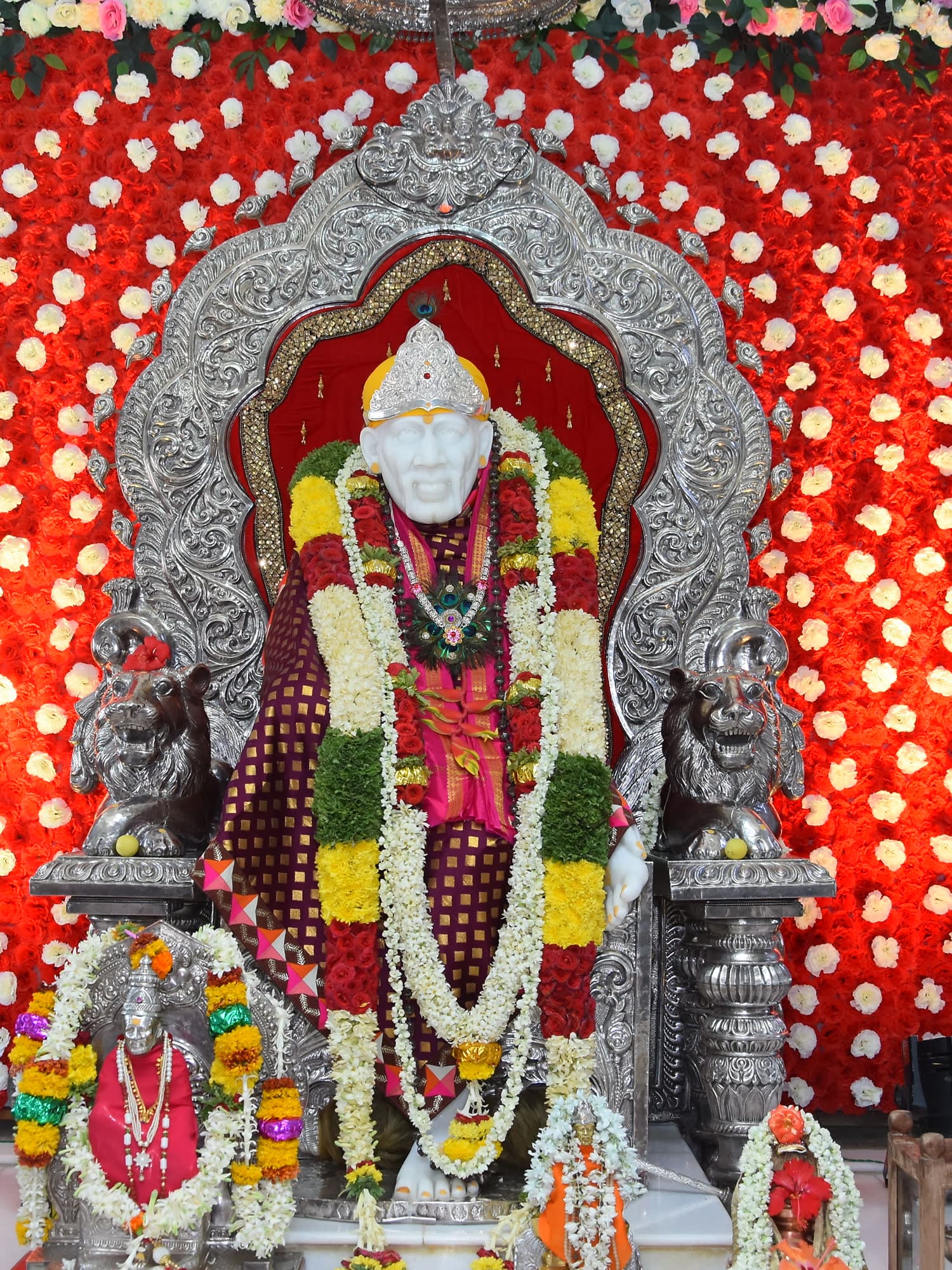 The murti dressed for the day, seated before a deep red backdrop hung with sweeping arcs of white and cream flowers.