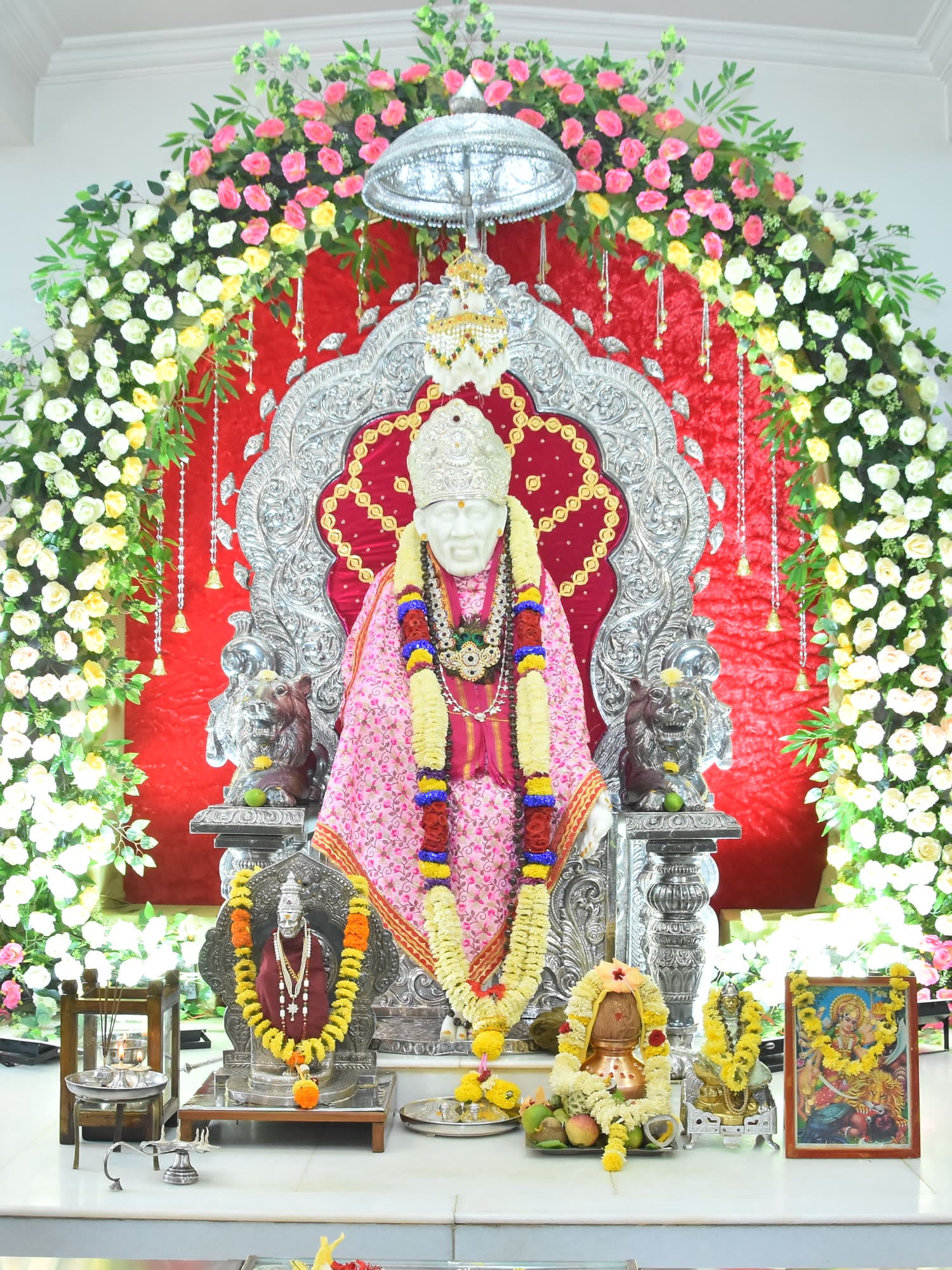 The murti in a pink robe beneath a canopy of rose and white flowers, with strands of blossom hanging on either side of the silver surround.